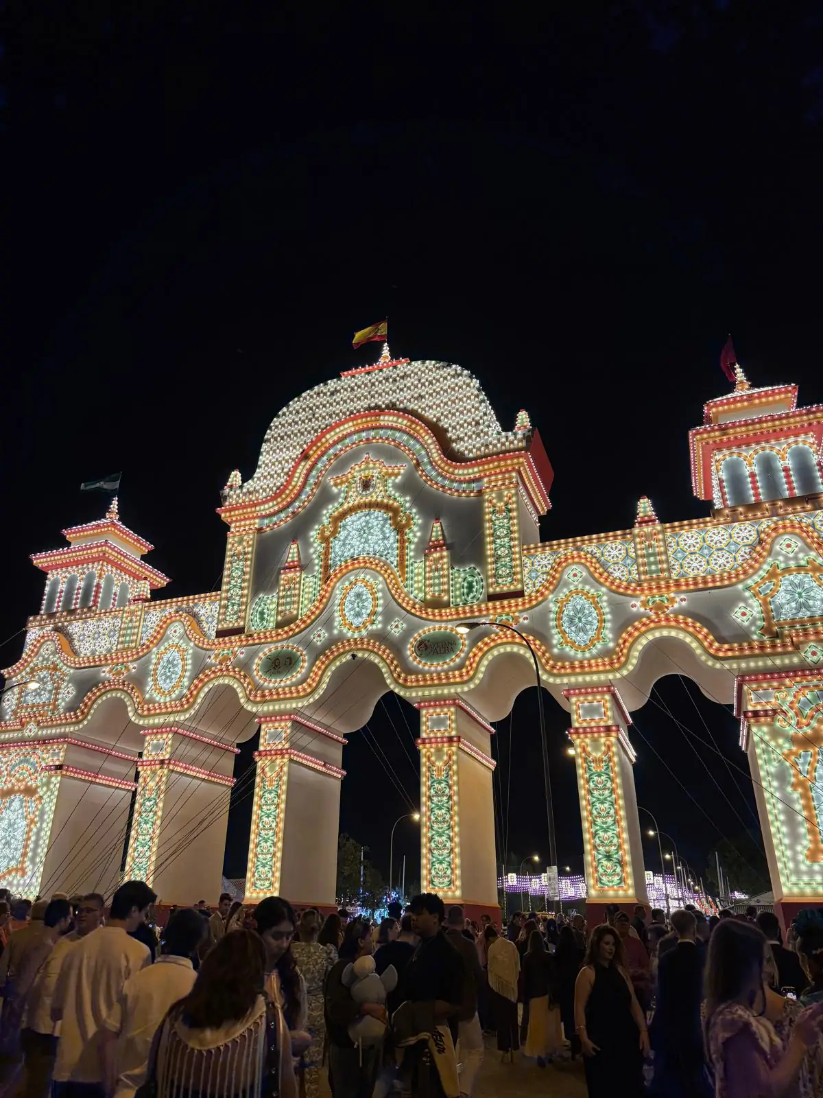 Portada iluminada da Feria de Abril, com visitantes à entrada do recinto, à noite