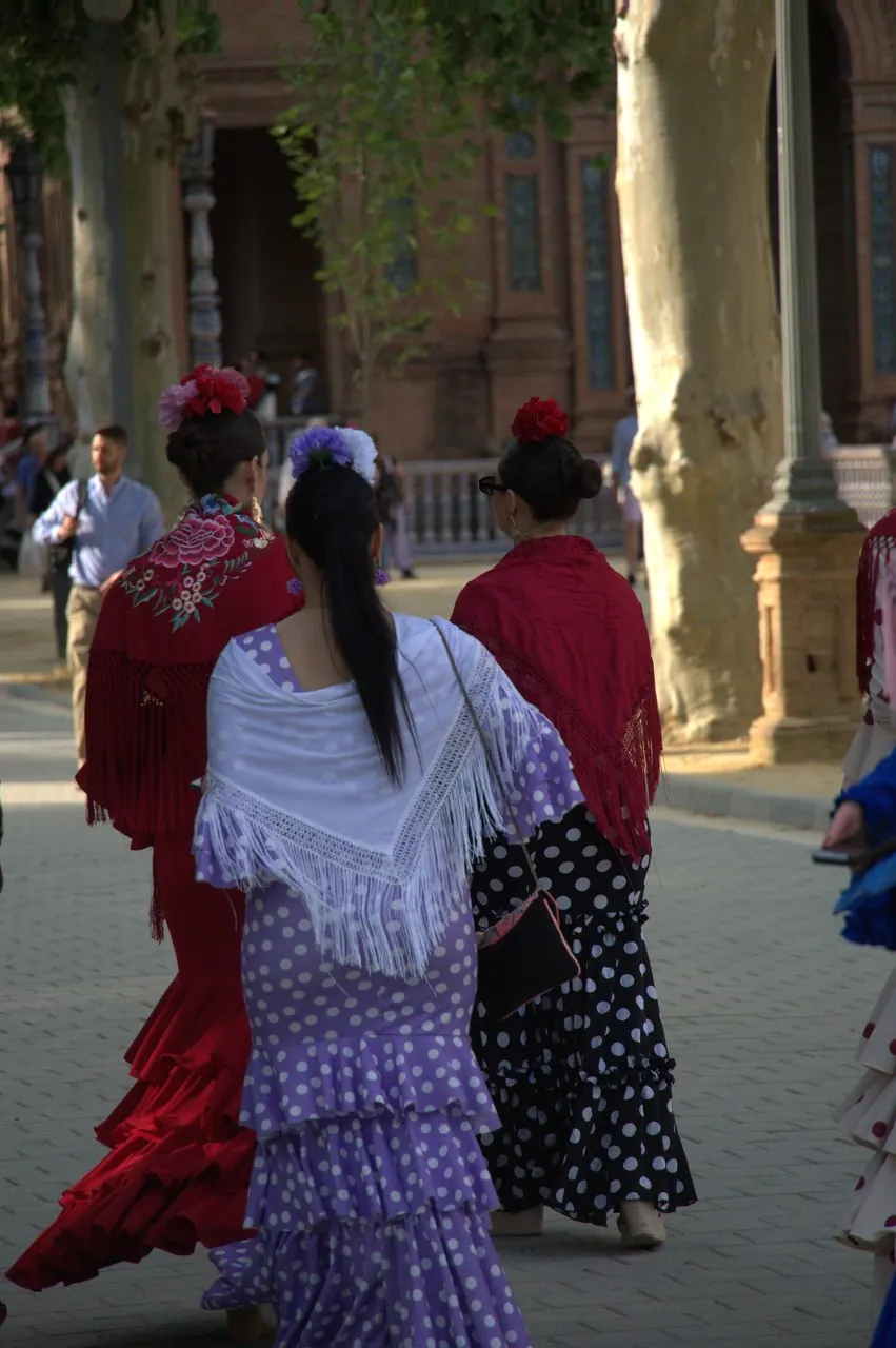 Visitantes com vestidos de flamenca e mantones, vistos de costas